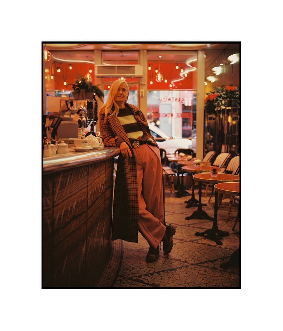 Blonde woman in a striped sweater and plaid coat posing in a vintage-style restaurant with red lighting and tables.