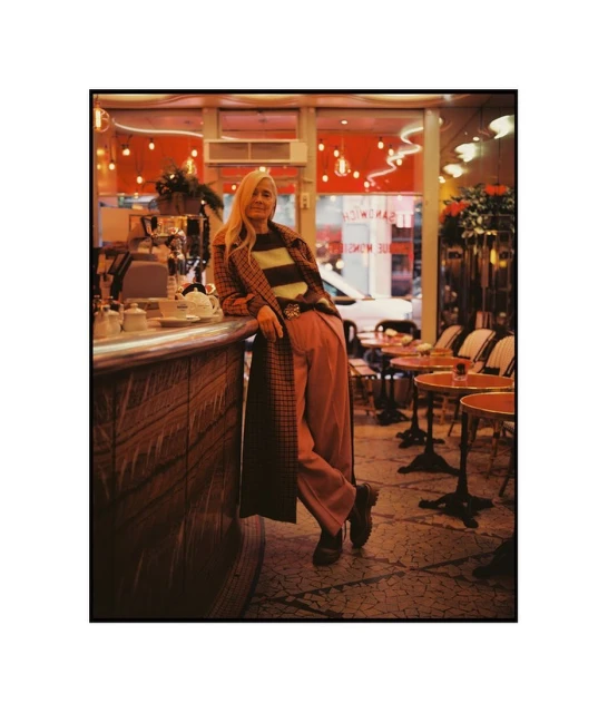 Blonde woman in a striped sweater and plaid coat posing in a vintage-style restaurant with red lighting and tables.