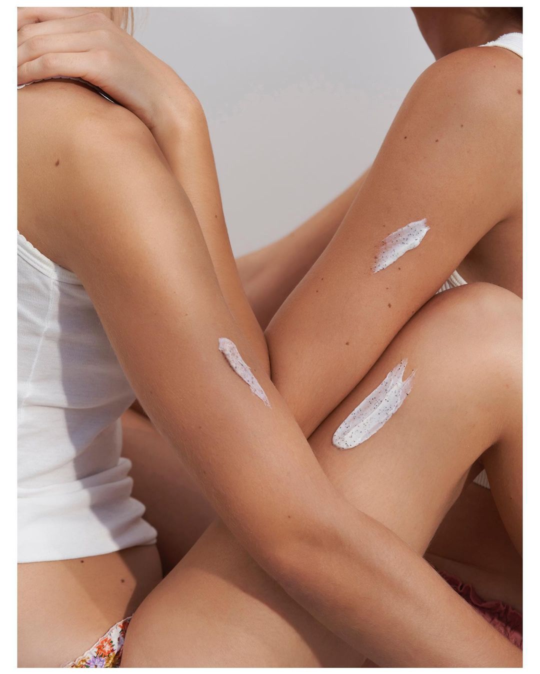 Close-up portrait of two women wearing white tank tops with exfoliating cream smeared on their arms, photographed by Eleonore Wismes