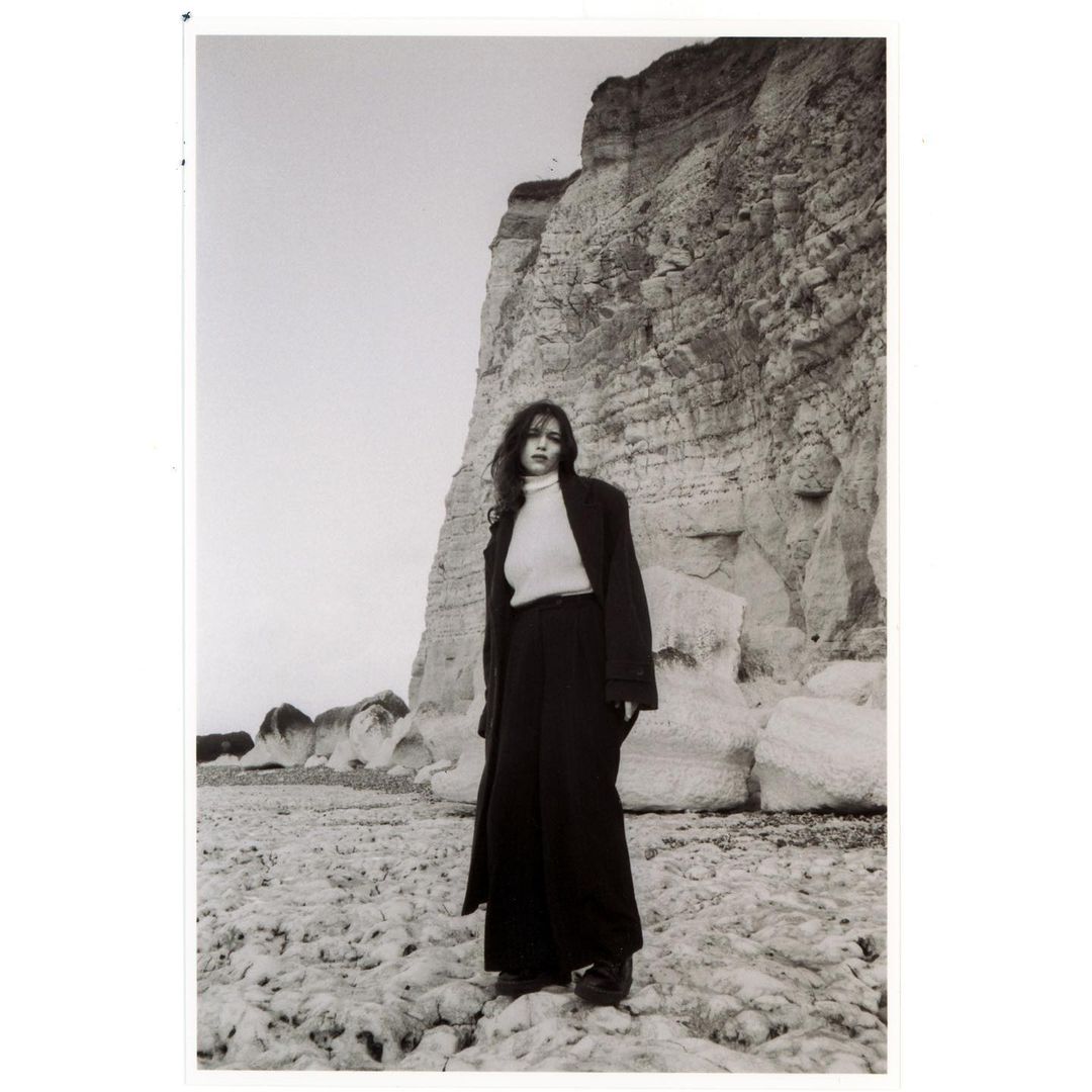 A monochrome portrait of a woman in a coat and pants standing on a rocky beach with a large cliff in the background and overcast sky.