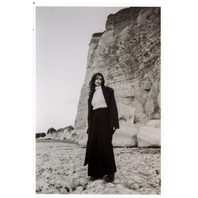 A monochrome portrait of a woman in a coat and pants standing on a rocky beach with a large cliff in the background and overcast sky.