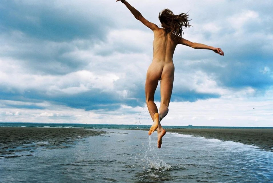 Woman leaps over water on a beach under a cloudy sky. The woman's arms are outstretched and water splashes around her feet.