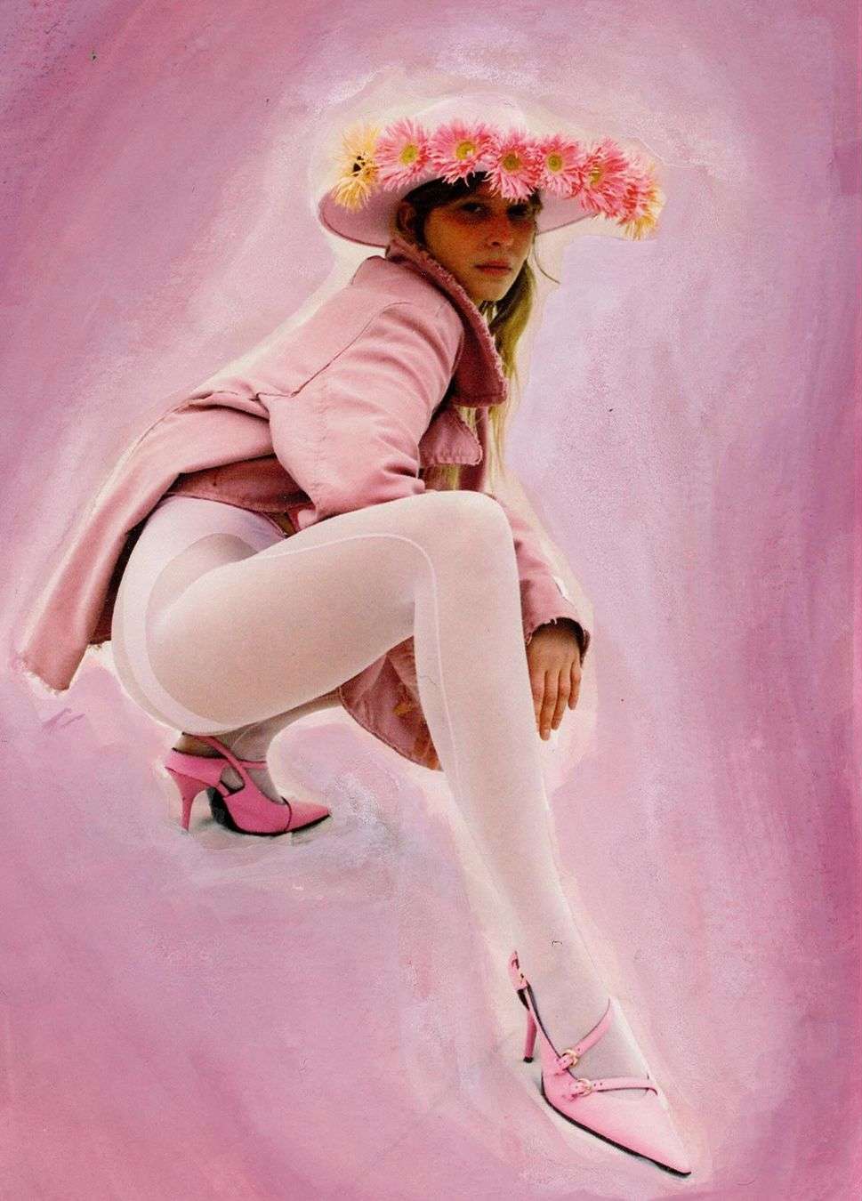 Woman in pink jacket, white tights, and pink heels poses with a flower hat against a pink painted backdrop, fashion-forward and stylish.