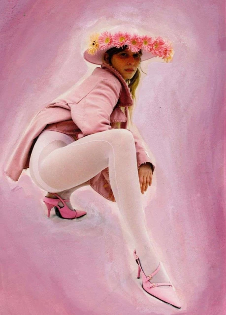 Woman in pink jacket, white tights, and pink heels poses with a flower hat against a pink painted backdrop, fashion-forward and stylish.