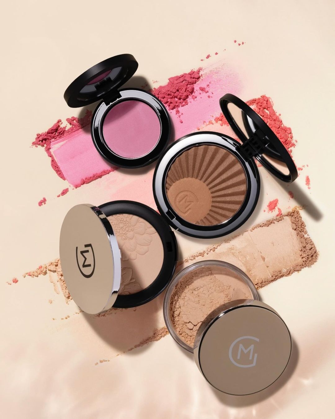 Overhead view of blush, bronzer, and powder makeup compacts in neutral and pink tones against a soft cream background with cosmetic powder.