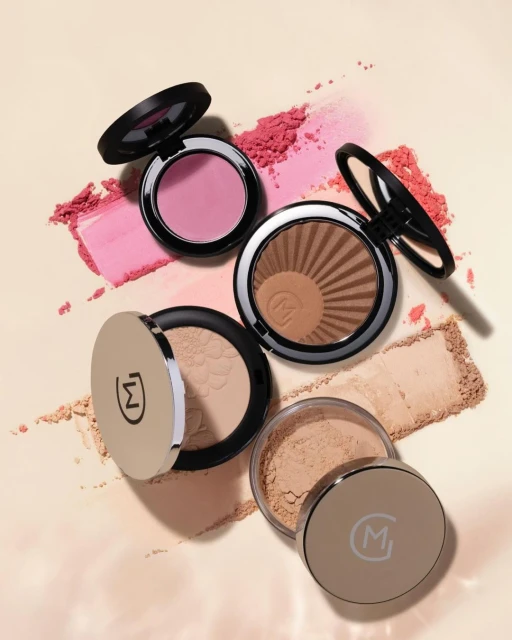 Overhead view of blush, bronzer, and powder makeup compacts in neutral and pink tones against a soft cream background with cosmetic powder.