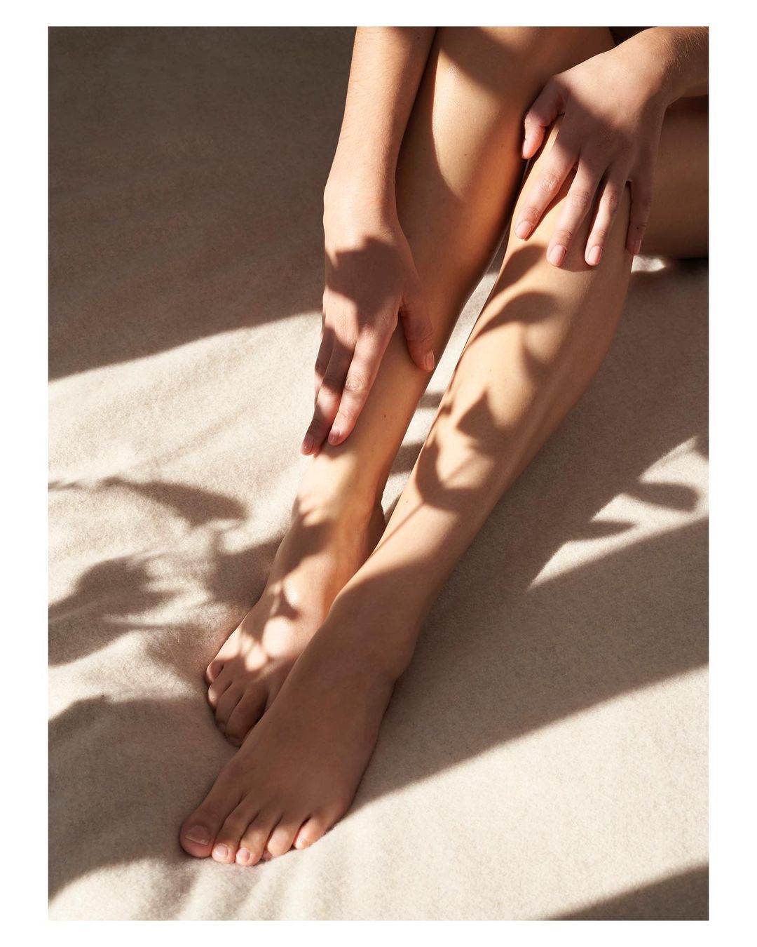 Close-up of an adult female seated with her legs extended, her hands resting on her legs, and shadows of foliage on the surface.