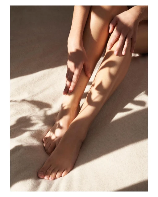Close-up of an adult female seated with her legs extended, her hands resting on her legs, and shadows of foliage on the surface.
