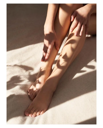 Close-up of an adult female seated with her legs extended, her hands resting on her legs, and shadows of foliage on the surface.