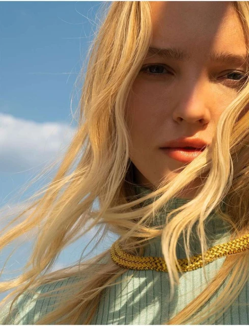 Close-up shot of a woman with long blonde wavy hair and a gold necklace, against a clear blue sky during daylight hours.