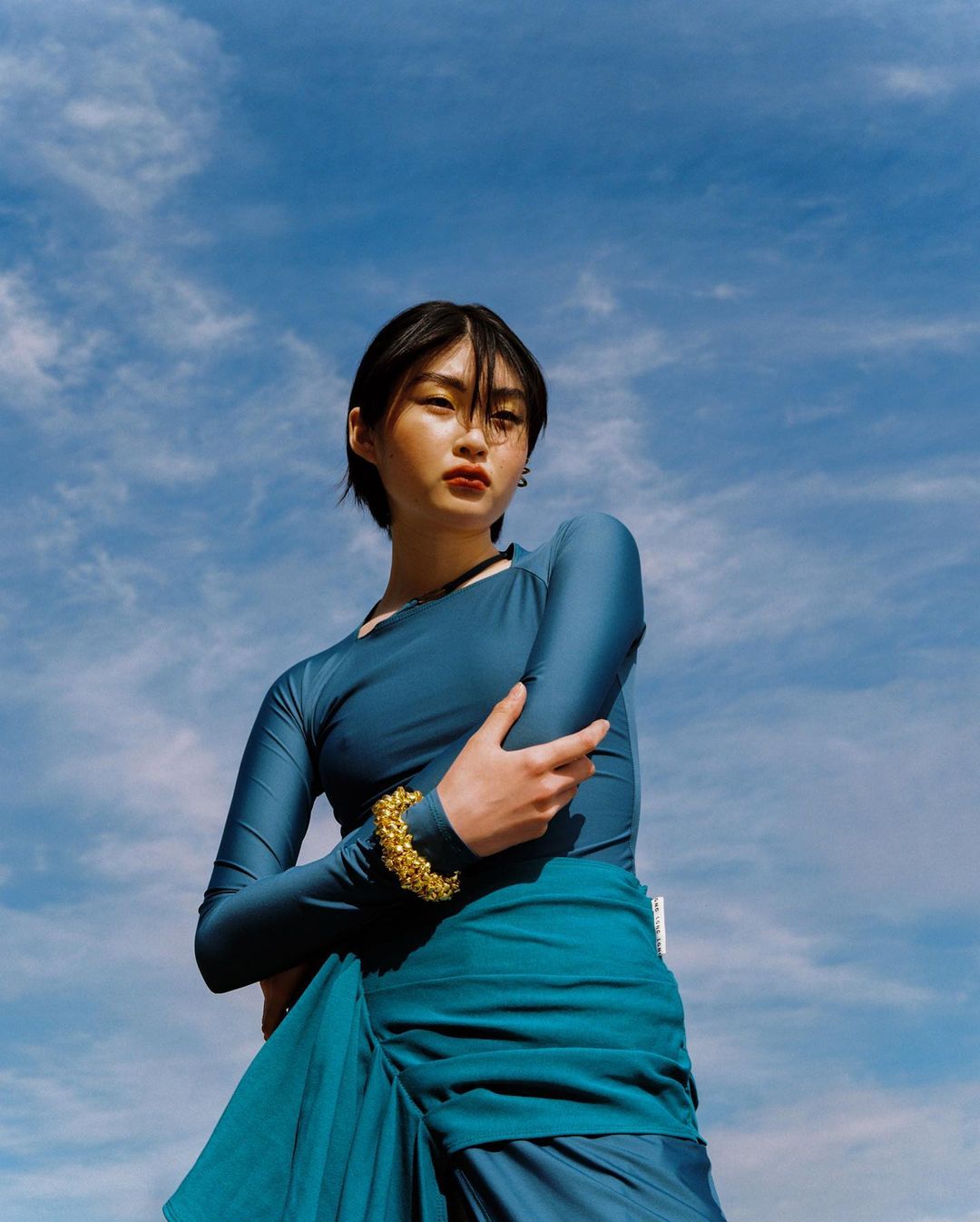Model in blue long-sleeve top wearing a gold textured bracelet against a bright sky background by Eleonore Wismes