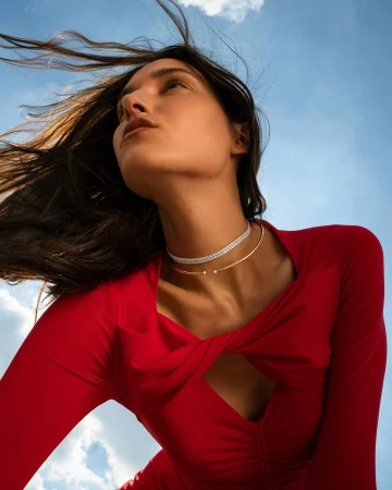 Low-angle shot of a woman in a bright red dress wearing choker necklaces against a blue sky with clouds, photographed by Eleonore Wismes.