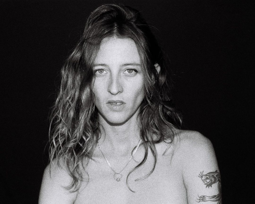 Black and white portrait of an adult woman with wavy shoulder-length hair, wearing a necklace, and featuring a tattoo on her arm.