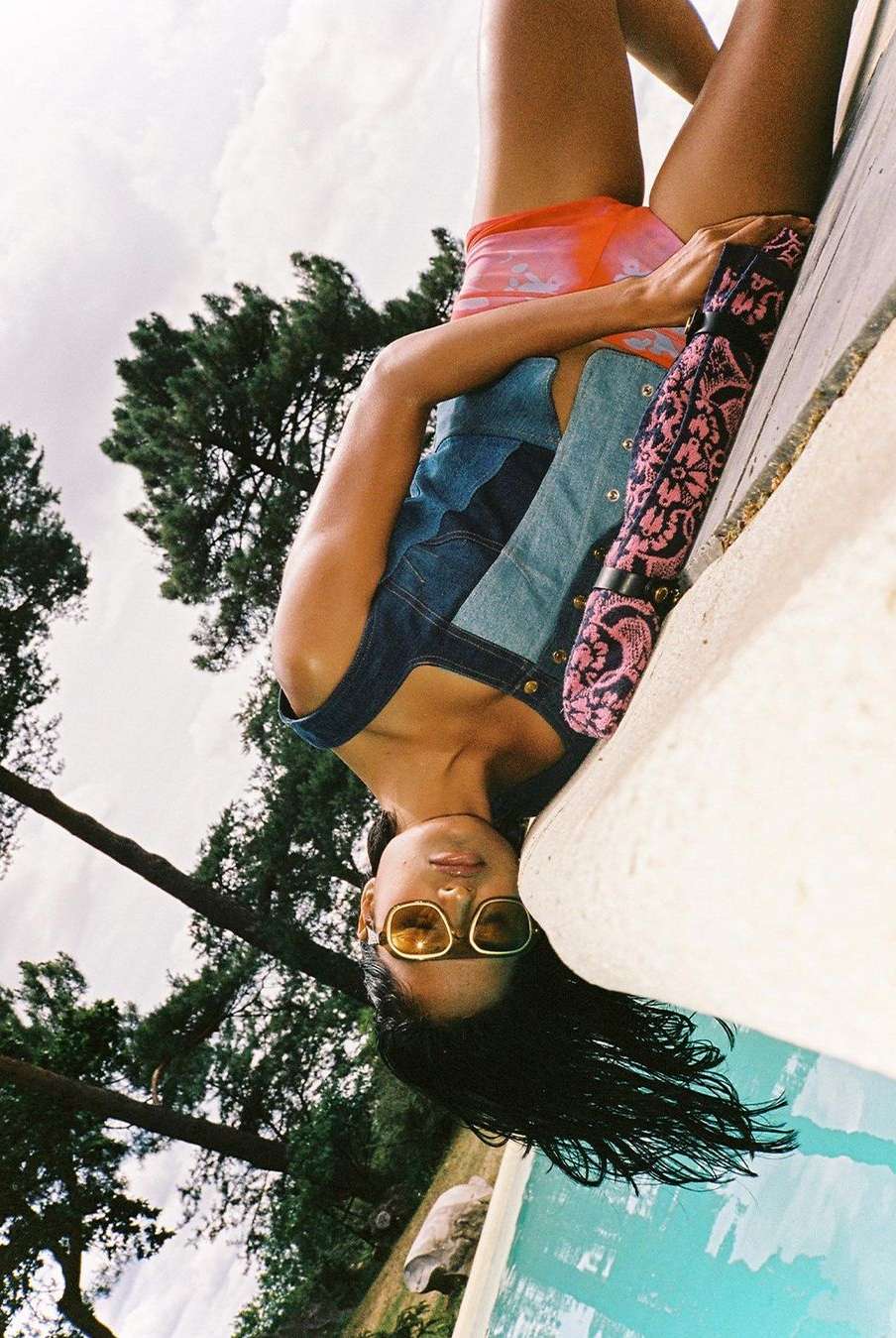 Woman in denim and swimwear poses near pool with trees and sky in background. Shot from high angle, inverted perspective for creative effect.