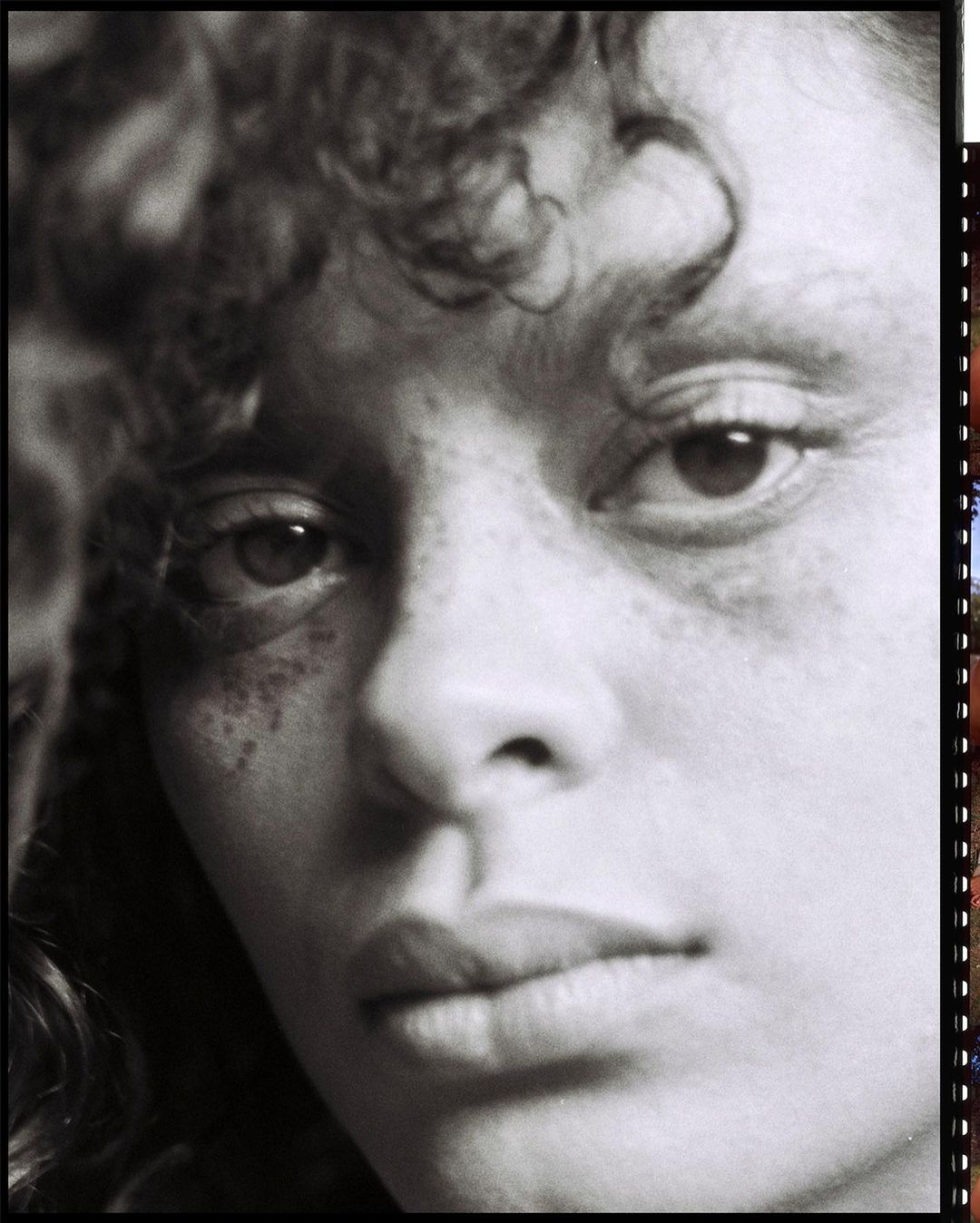 Close-up black and white portrait of a woman with curly hair, freckles, and natural makeup, capturing an intimate and raw aesthetic.