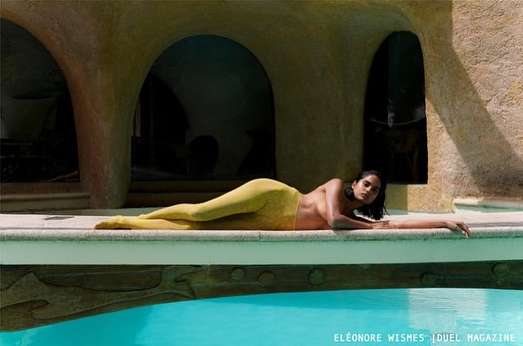 Adult woman with tan skin poses in yellow leggings, lying on a ledge beside an outdoor pool, with a textured arched building behind.