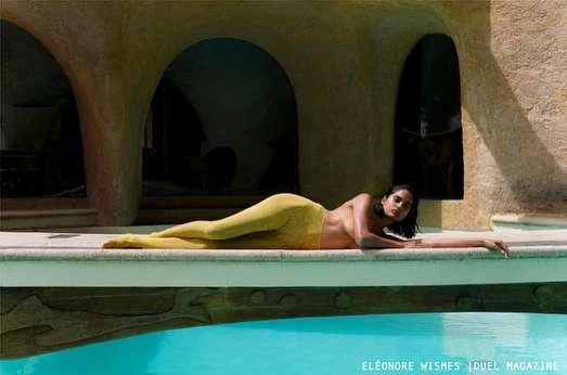 Adult woman with tan skin poses in yellow leggings, lying on a ledge beside an outdoor pool, with a textured arched building behind.