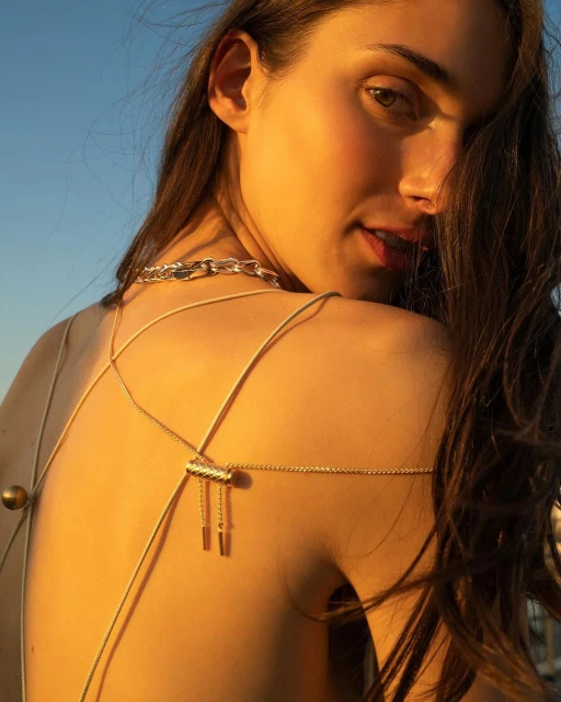 Young woman with long brunette hair looks over her shoulder, illuminated by golden sunlight, wearing delicate jewelry and a chain choker.
