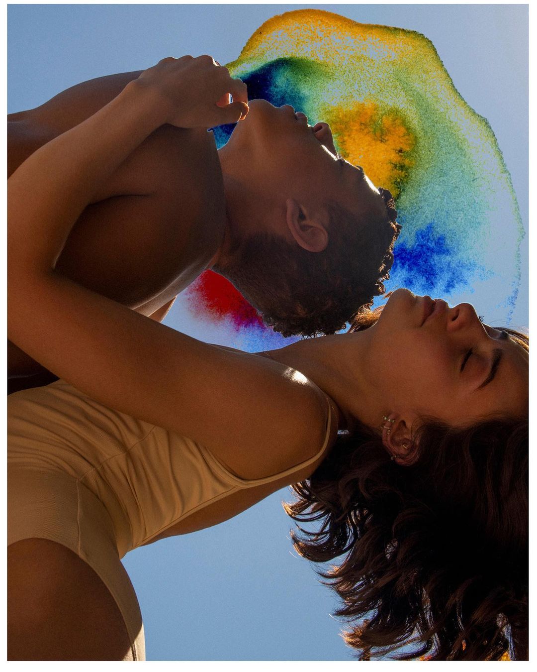 Eye-level shot of a shirtless man embracing a woman in a swimsuit, facing up to the sky with colorful paint-like effects above them.