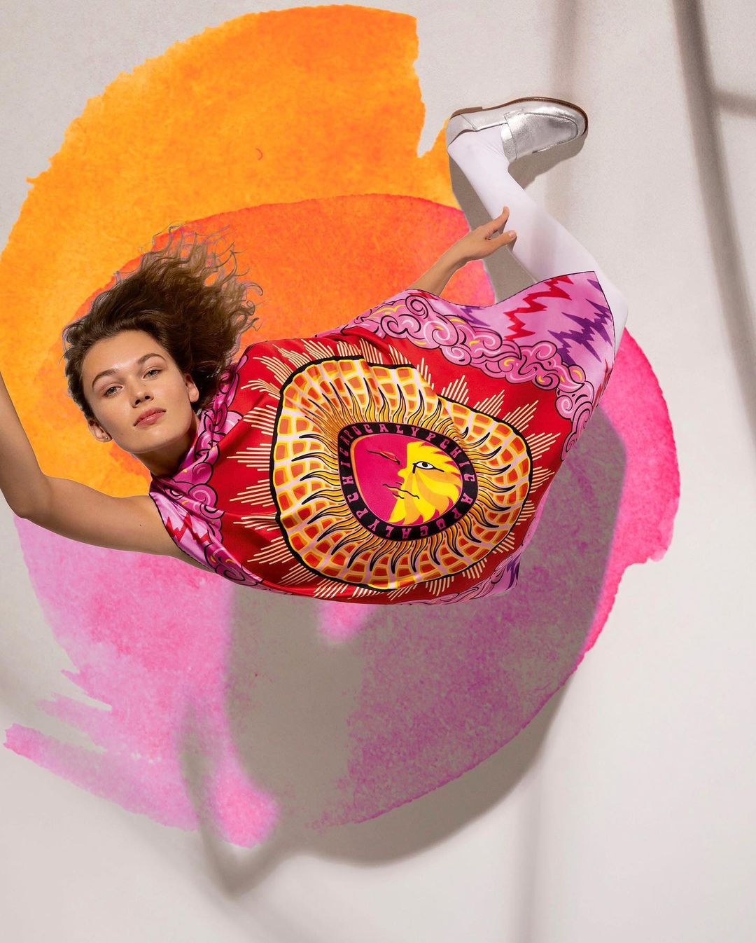 Woman in a colorful dress poses in mid-air against a backdrop of blended orange and pink circular watercolor paint strokes.