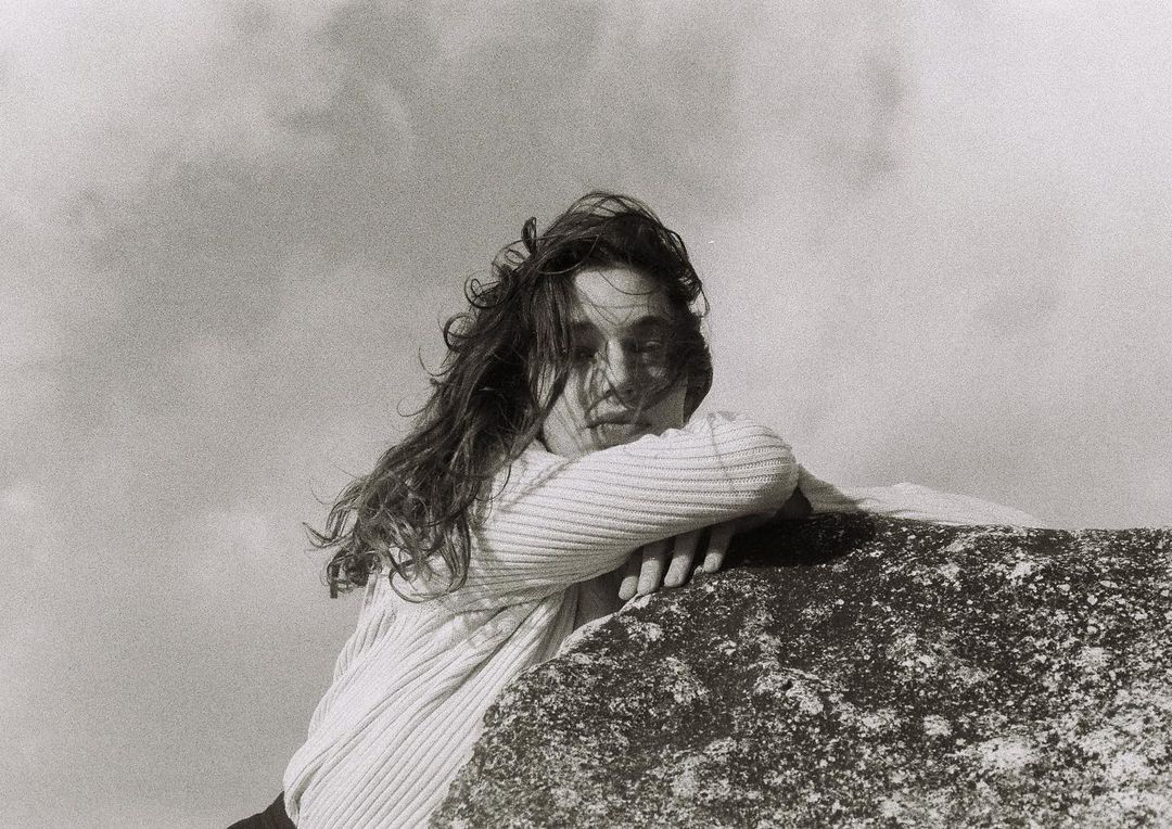 Monochrome portrait of a woman with long, wavy hair, leaning on a textured rock formation against a cloudy sky background.