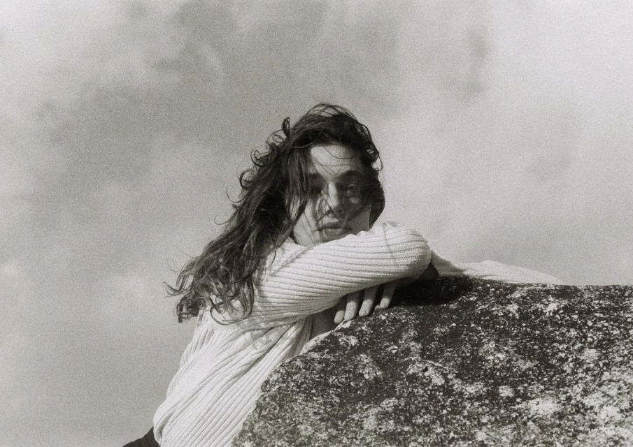 Monochrome portrait of a woman with long, wavy hair, leaning on a textured rock formation against a cloudy sky background.