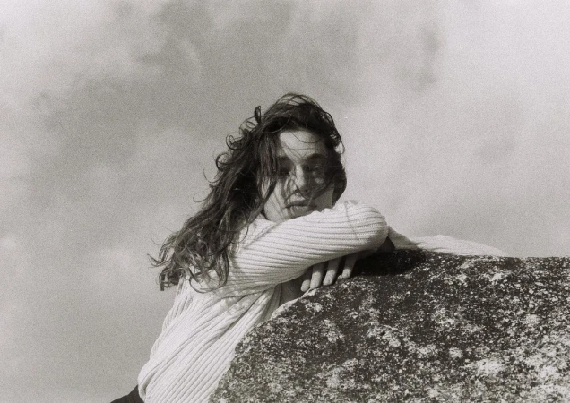 Monochrome portrait of a woman with long, wavy hair, leaning on a textured rock formation against a cloudy sky background.