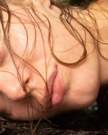 Close-up portrait of a woman with wet brown hair framing her face and lips resting on a dark surface