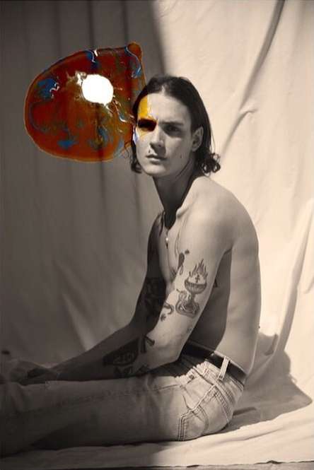 Young man with tattoos and a yellow painted face sits near a large, colorful abstract shape in front of a white draped background.