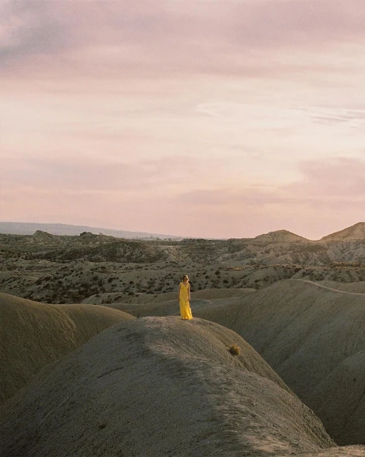 Woman in long yellow dress stands on a tan desert dune landscape under a pastel sky at sunset, evoking a sense of solitude.
