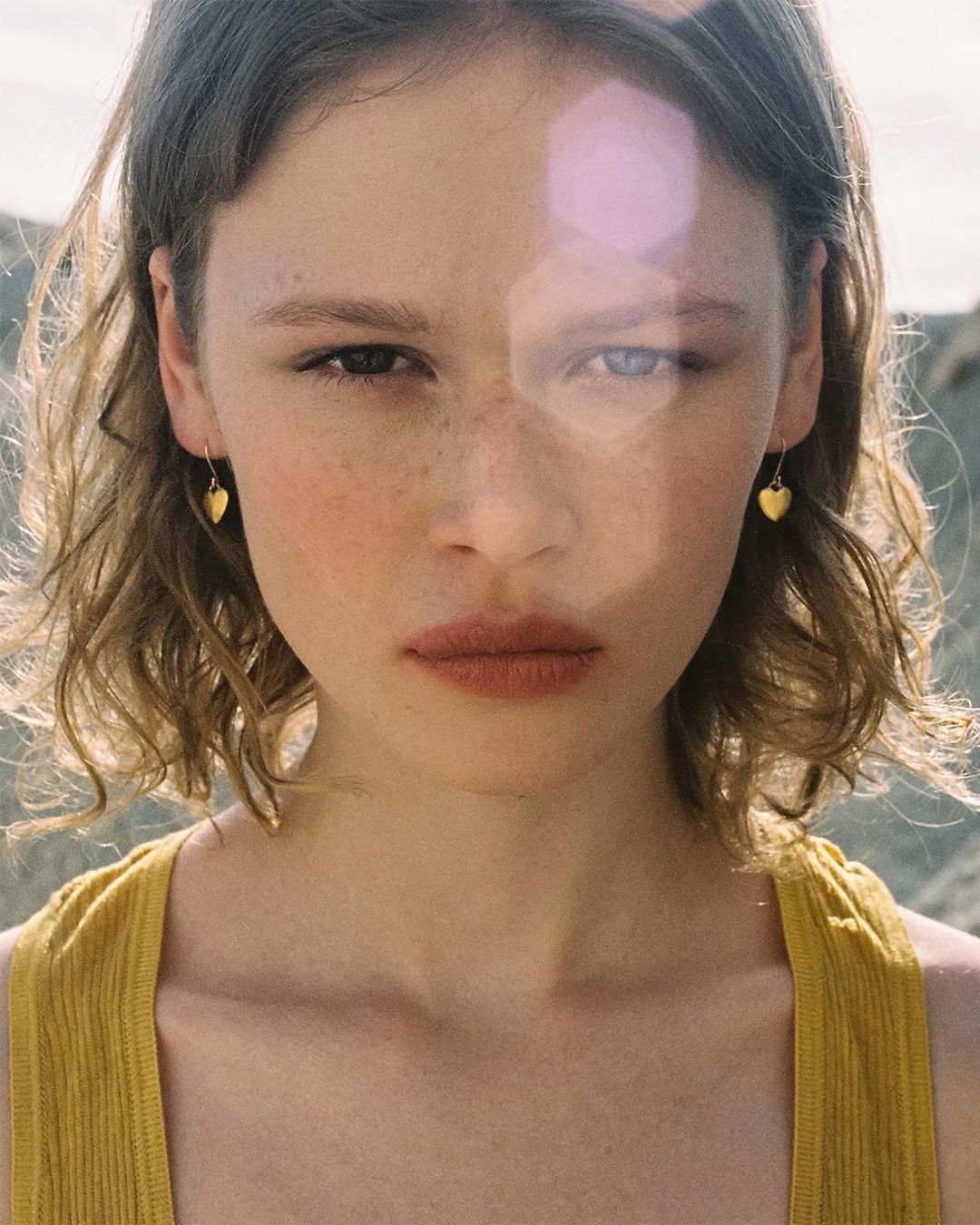 Close-up of a young woman with wavy light brown hair, wearing a yellow tank top and small gold heart earrings, against an outdoor background.