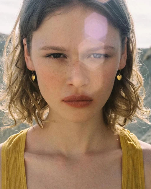 Close-up of a young woman with wavy light brown hair, wearing a yellow tank top and small gold heart earrings, against an outdoor background.