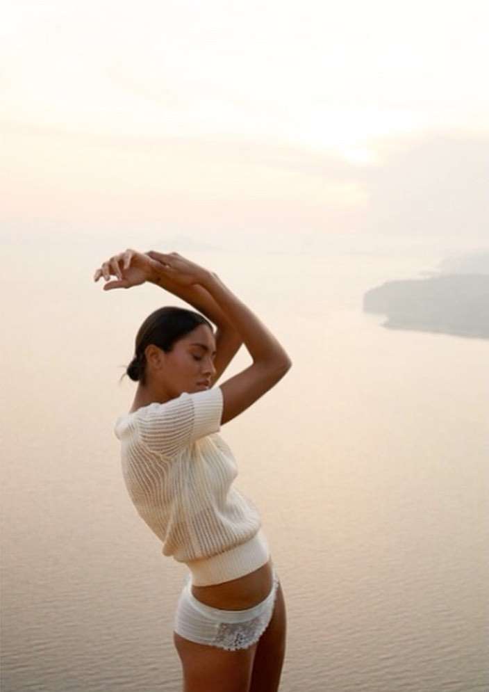 Adult woman in a white knit top and underwear poses outdoors with her arms raised above her head and a body of water in background.