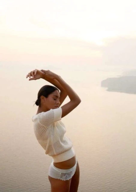 Adult woman in a white knit top and underwear poses outdoors with her arms raised above her head and a body of water in background.