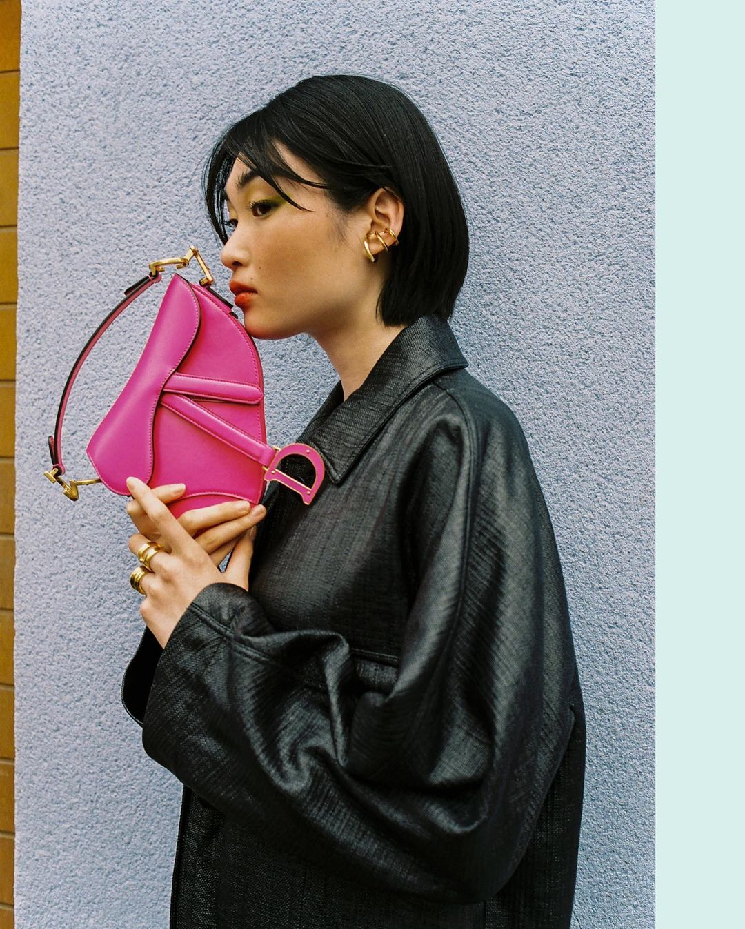 Profile of a woman in a black coat holding a bright pink Dior purse against a purple wall with yellow trim, showcasing fashion and style.