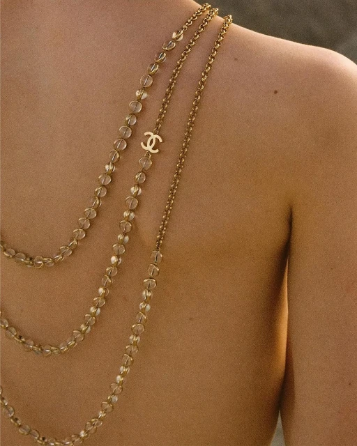 Close-up studio shot of a bare-backed adult female wearing three layered gold Chanel necklaces against a blurred tan background.