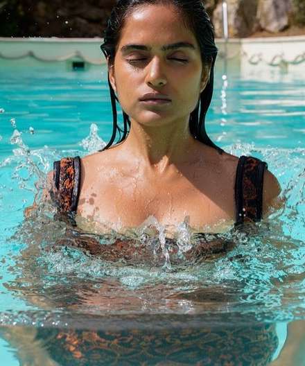 Woman with closed eyes in patterned bathing suit emerges from a swimming pool, water droplets glistening on her skin. Sunlit, outdoor setting.
