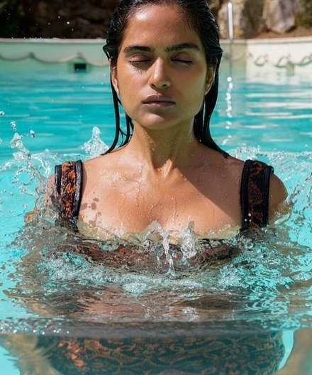 Woman with closed eyes in patterned bathing suit emerges from a swimming pool, water droplets glistening on her skin. Sunlit, outdoor setting.