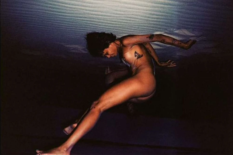 Nude adult female with short dark hair posing under a light source, with tattoos on chest and arm, against a dark background.