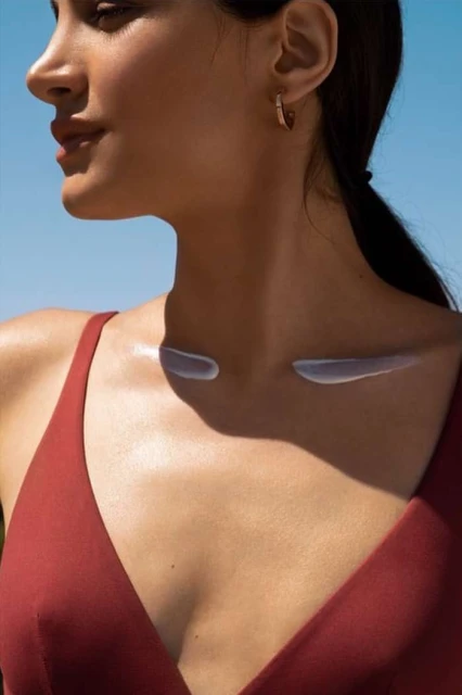 Close-up shot of a woman in a red dress with cream on her collarbones against a clear blue sky, highlighting skincare and summer.