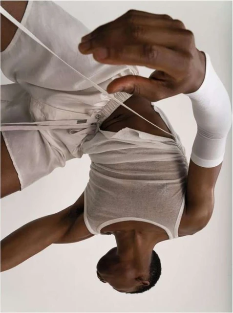 An athletic Black male in a casual white outfit poses in an upside-down orientation on a white backdrop, holding white string.