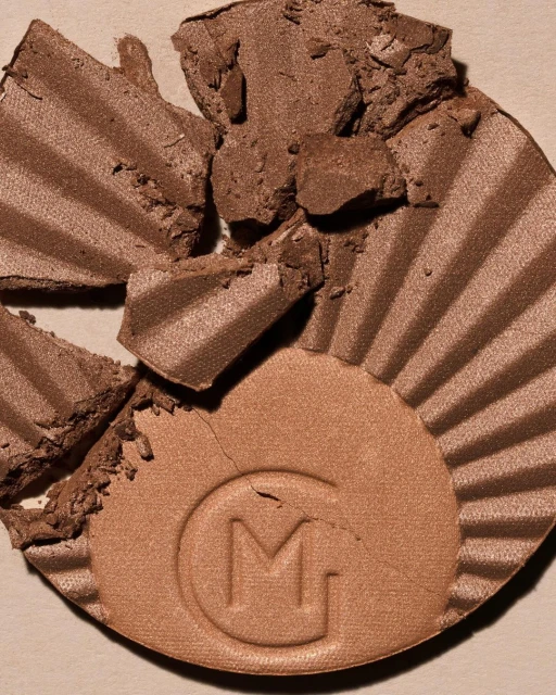 Close-up of a broken bronze cosmetic compact with a radial pattern and a textured letter 'M' on a beige background.