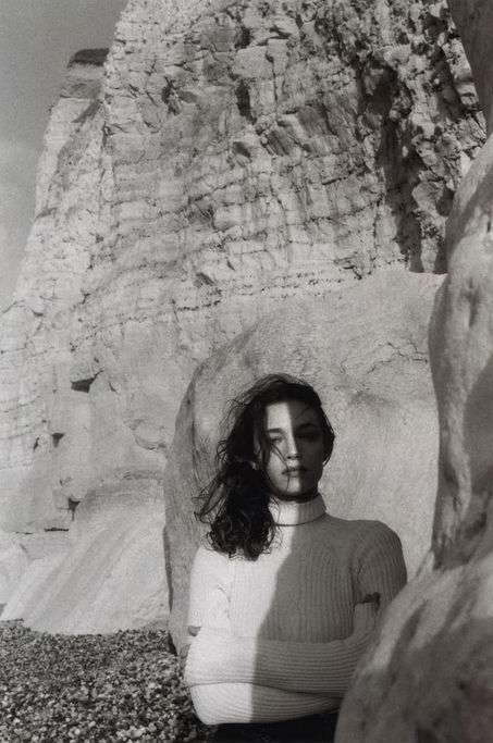 Monochrome portrait of a young woman with windblown hair, wearing a turtleneck sweater, against a rocky coastal background with shadows.