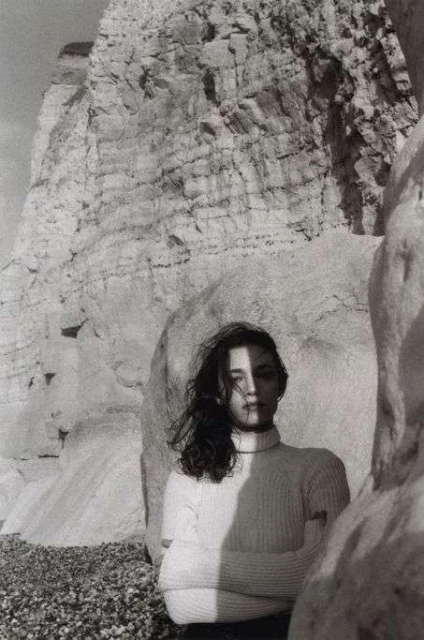 Monochrome portrait of a young woman with windblown hair, wearing a turtleneck sweater, against a rocky coastal background with shadows.