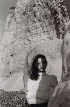 Monochrome portrait of a young woman with windblown hair, wearing a turtleneck sweater, against a rocky coastal background with shadows.