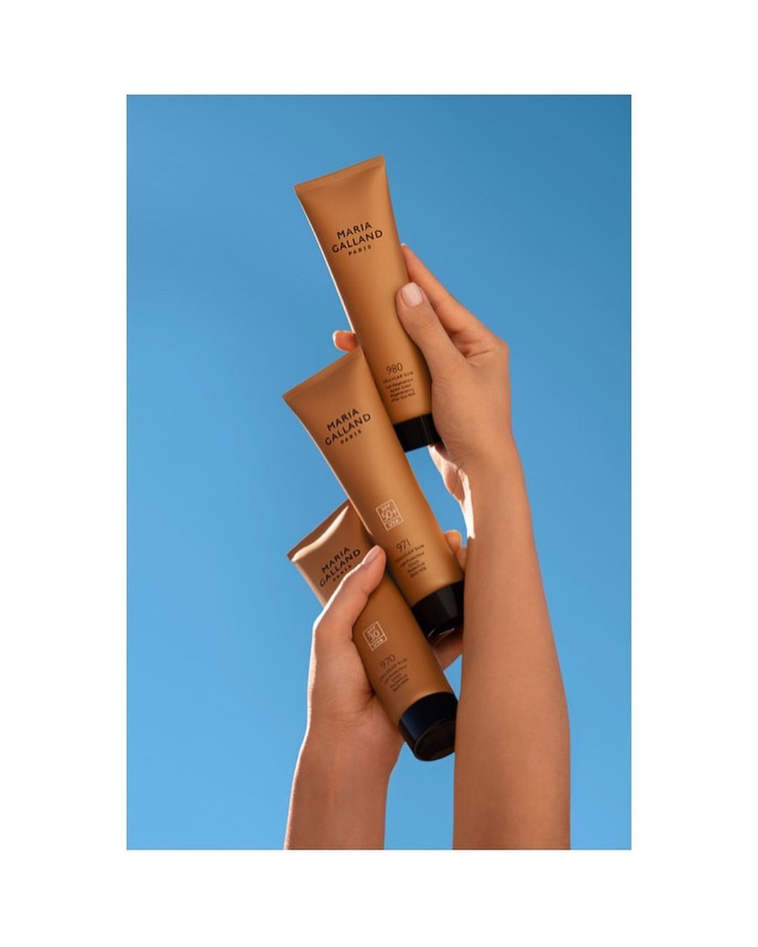 Trio of tanning lotion tubes with black lids held by light-skinned woman's hands against a bright blue sky background in studio shot.