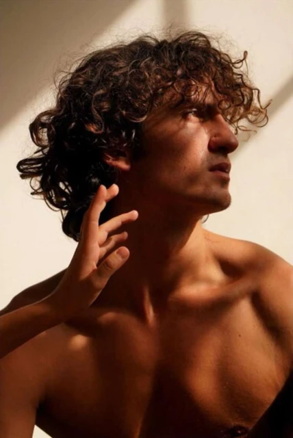 Studio portrait of a bare-chested man with curly hair, his face lit by warm, natural light with a woman's hand gently touching his face.