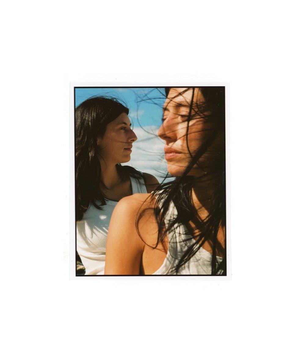 Close-up of two women with dark hair and white tops against a blue sky, one in profile and the other partially visible in the background.