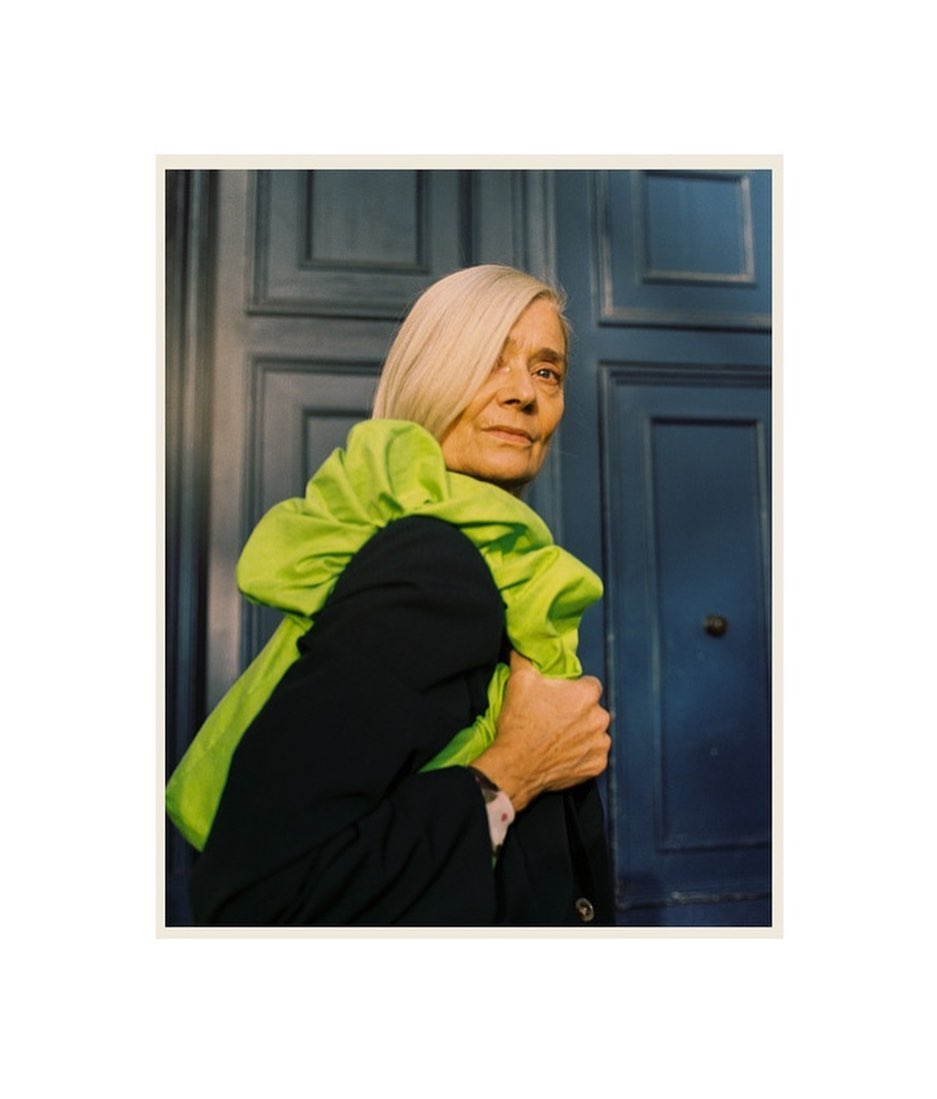 Older adult woman in a black jacket poses indoors, wearing a bright green scarf against a dark blue paneled door backdrop.