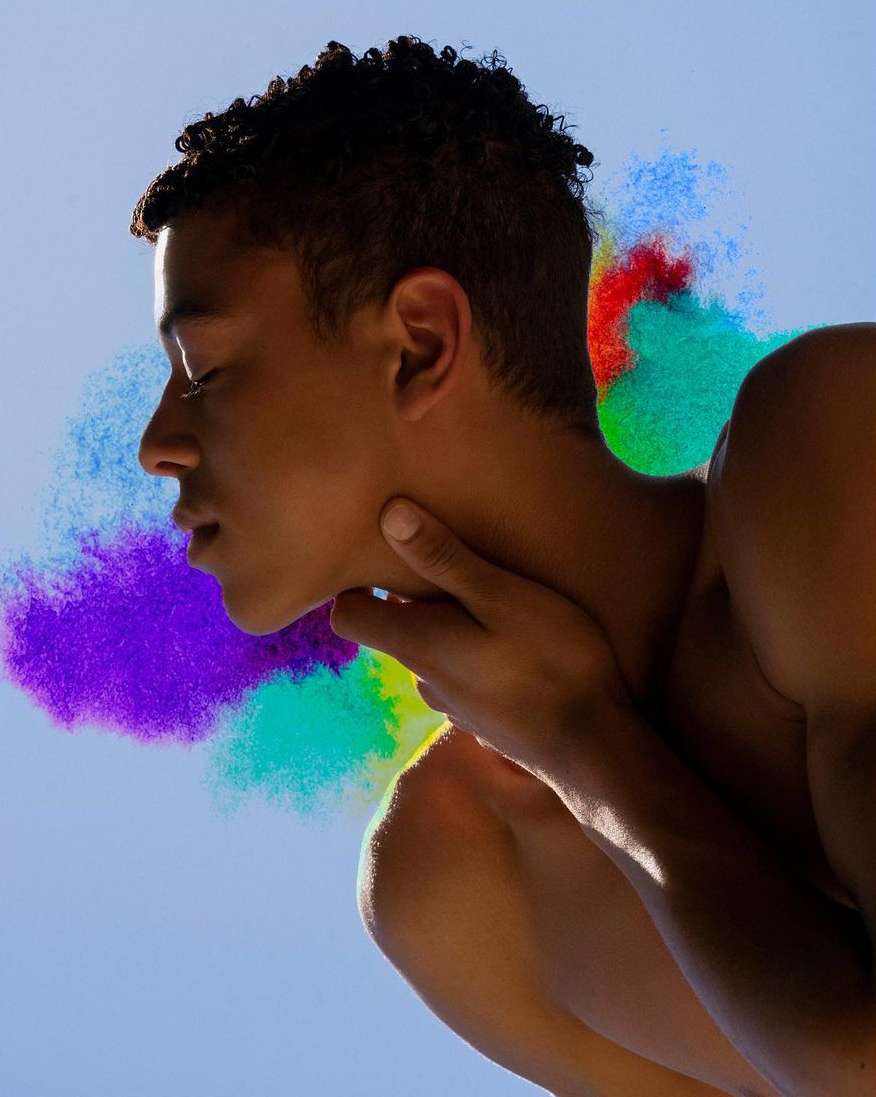 Portrait of a shirtless adult with dark curly hair against a blue background with red, green, purple, and blue powder paint.